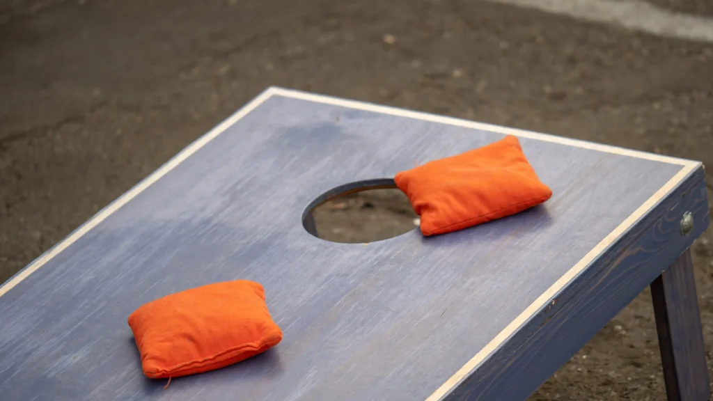 A blue wooden cornhole board with two orange cornhole bags thrown near the hole, placed on a paved surface