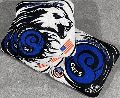 Two cornhole bags are stacked on a gray surface, both featuring a white tiger head with blue eyes and black accents on a black and white background. The top bag clearly shows the tiger head with "TYPHOON" above it. Below the tiger is a large blue "S" with "CAT 5" and "UNITED WE THROW" written around it. Small American flag patches are visible. The bottom bag is partially obscured but shows similar design elements, including the blue "S," "CAT 5," and text like "CORNHOLE SCENARIO." A small circular logo with "PIR" and an American flag design is also visible.
