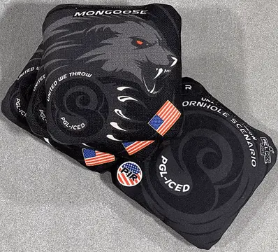 Two black cornhole bags are shown on a gray surface. The top bag features a large, menacing gray mongoose head with glowing red eyes and bared fangs. "MONGOOSE" is written above the head. Below the mongoose, there's a dark gray swirling design with "UNITED WE THROW" and "PGL-ICED" visible, along with multiple small American flag patches. The bottom bag is mostly black with a similar dark gray swirling pattern and text that includes "CORNHOLE SCENARIO" and "PGL-ICED." A small circular patch with "PIR" is also visible on the bottom bag.