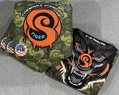 Two cornhole bags with a camouflage pattern are visible on a gray surface. The top bag is primarily olive green with an orange circular logo featuring a stylized "S" and "TIGER" written below it. Above the logo, "CORNHOLE SCENARIO" is written in a semi-circle. To the left, there are three small circular patches with red and blue designs. The bottom bag is mostly black with a ferocious tiger's face design. The tiger has blue eyes and orange accents. A smaller "CORNHOLE SCENARIO" text and an "S" logo are also present within the tiger design.