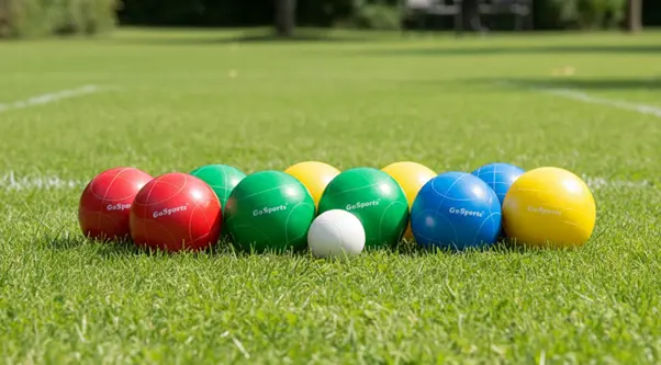 A set of colorful bocce balls—red, green, blue, and yellow—arranged in a row on a grassy lawn, with a small white pallino ball placed in front of them.