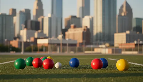 Colorful bocce balls spread across a turf field with a white pallino ball in front, set against a backdrop of tall city skyscrapers and an urban skyline.